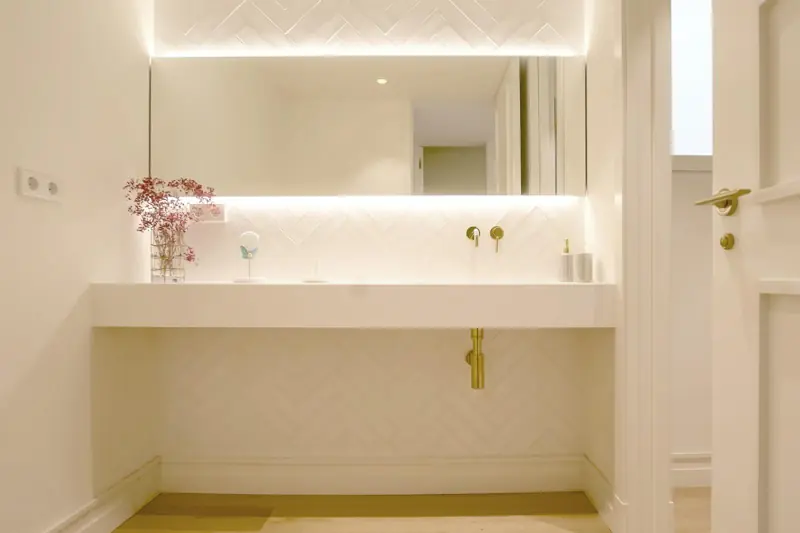 Baño con lavabo suspendido, grifería dorada e iluminación indirecta